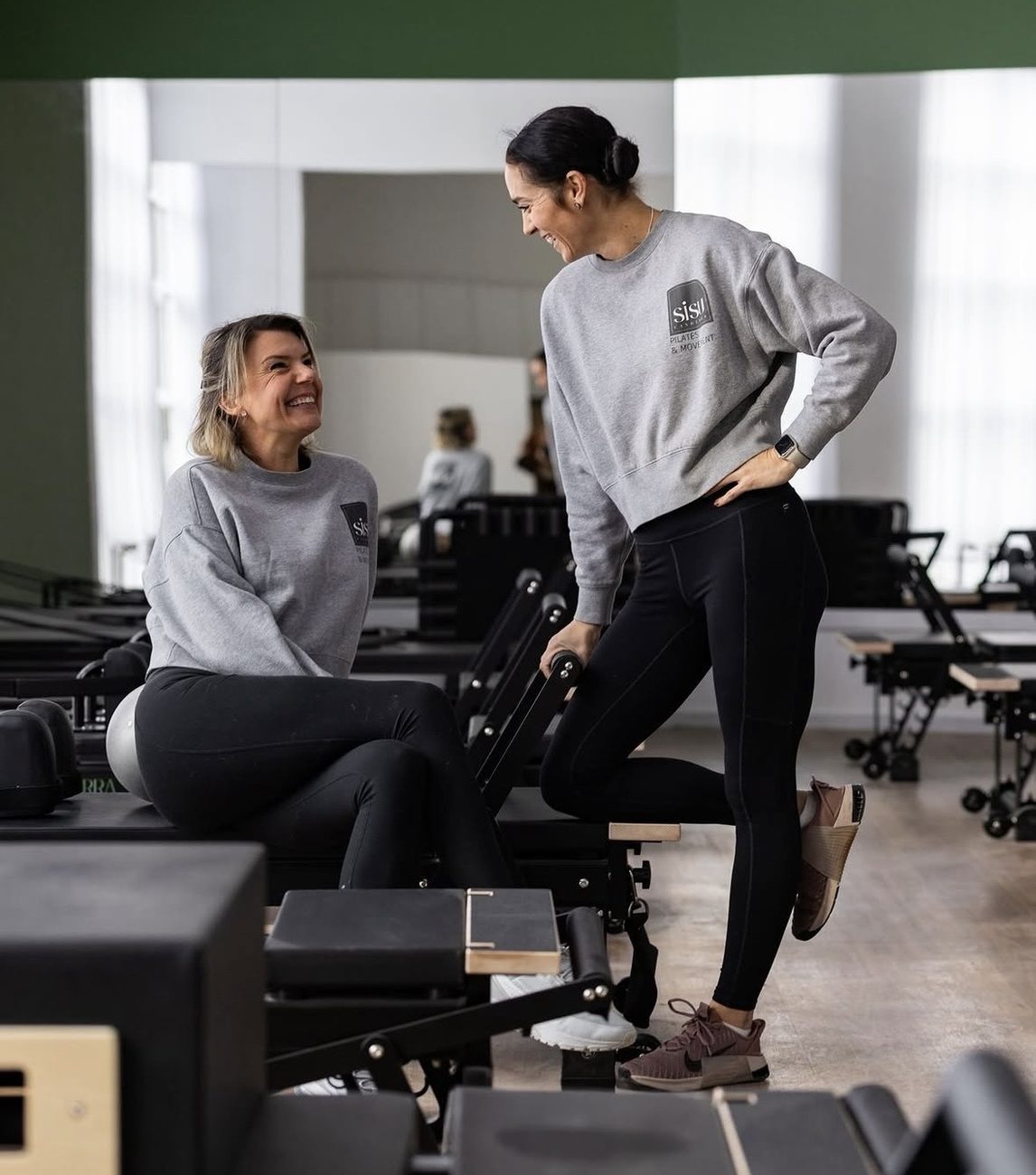 Two SiSU instructors laughing together in the reformer studio